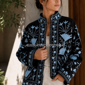 Pode incluir: Uma jaqueta preta com bordados florais em azul claro, gola alta e mangas compridas. O texto ASSUHANDICRAFTINDIA é visível no centro. A jaqueta é usada sobre uma blusa branca e calças brancas, criando um contraste de cores.