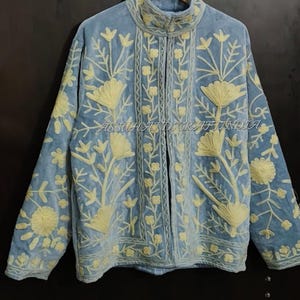 May include: A light blue jacket featuring cream-colored floral embroidery throughout the front and sleeves. The garment has a high collar, long sleeves, and a central opening. The text ASSUHANDICRAFTINDIA is visible across the center of the jacket.
