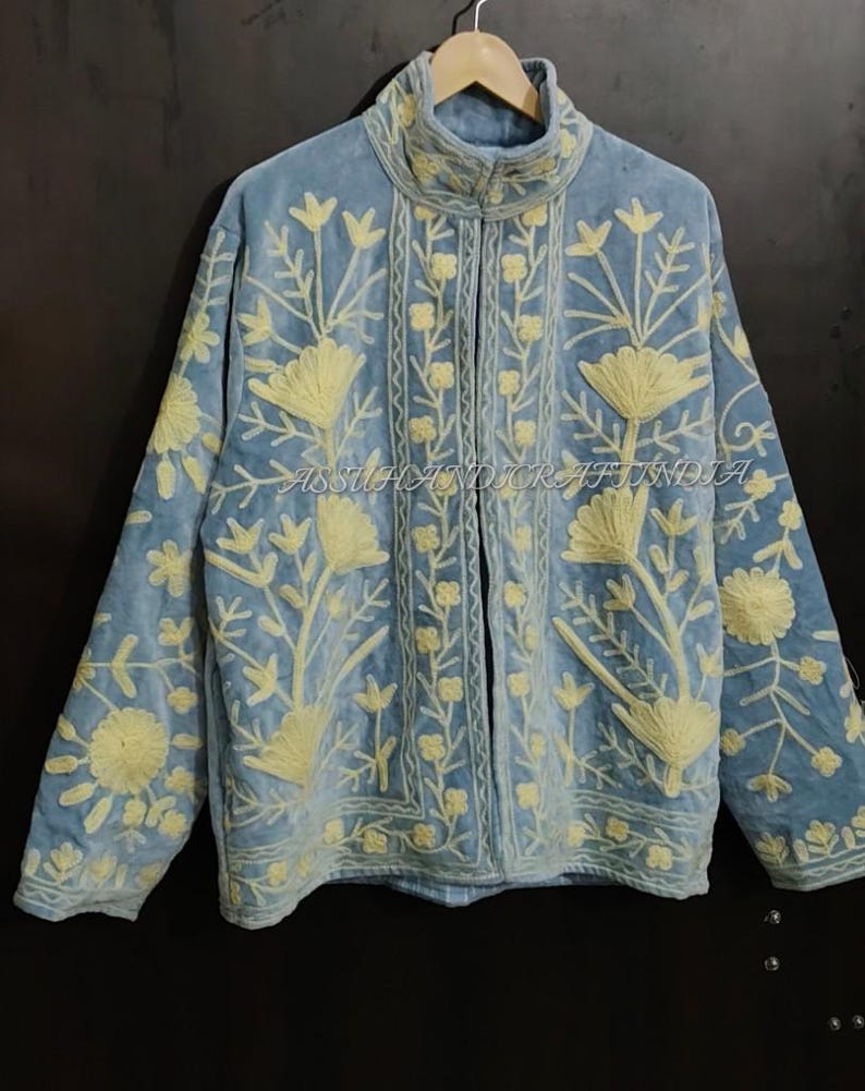 May include: A light blue jacket featuring cream-colored floral embroidery throughout the front and sleeves. The garment has a high collar, long sleeves, and a central opening. The text ASSUHANDICRAFTINDIA is visible across the center of the jacket.