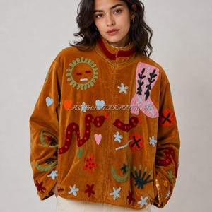 Pode incluir: Uma jaqueta de veludo amarelo-mostarda com gola alta e fecho de correr. A jaqueta é decorada com motivos bordados coloridos, incluindo um sol, uma bota rosa, cobras, corações, estrelas, flores e luas. O texto ASSHANDICRAFTINDIA é visível.