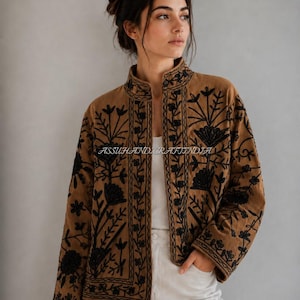 Puede incluir: Una chaqueta marrón con bordados florales negros, cuello alto y mangas largas. La prenda está abierta por delante, dejando ver una camiseta blanca y pantalones blancos. El texto ASSUHANDICRAFTINDIA es visible en la tela de la chaqueta.