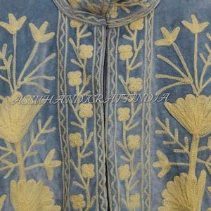 May include: A light blue velvet jacket featuring a high collar and intricate yellow floral embroidery. The garment displays vertical patterns of small flowers and large stylized blossoms. The text ASSUHANDICRAFTINDIA is visible across the center.
