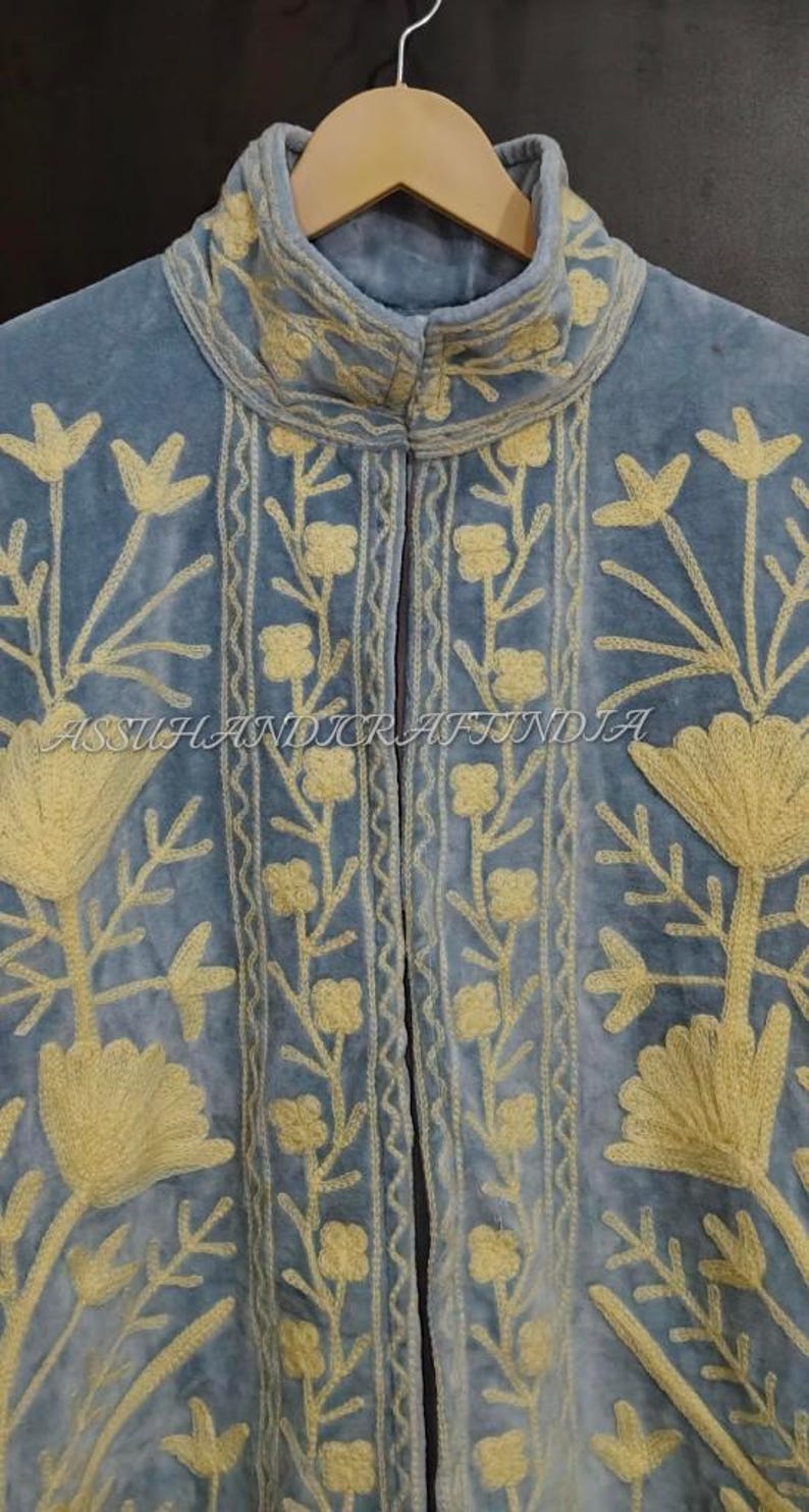 May include: A light blue velvet jacket featuring a high collar and intricate yellow floral embroidery. The garment displays vertical patterns of small flowers and large stylized blossoms. The text ASSUHANDICRAFTINDIA is visible across the center.