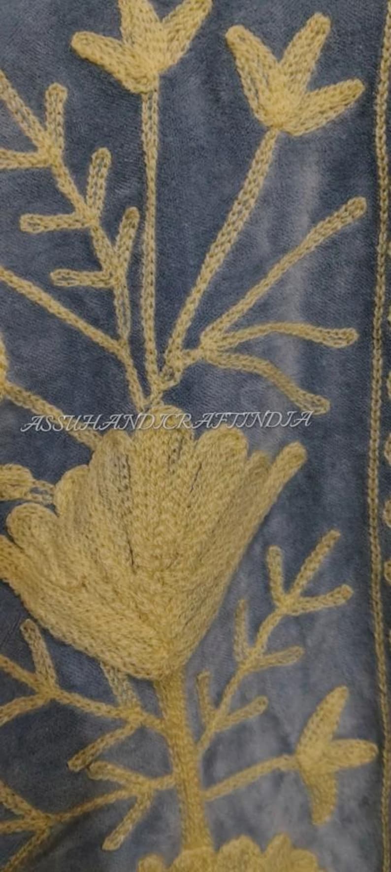 May include: A blue fabric featuring intricate yellow floral embroidery with stems and leaves. The text ASSUHANDICRAFTINDIA is embroidered in the center. The design displays a large central flower with smaller buds and foliage extending upwards.