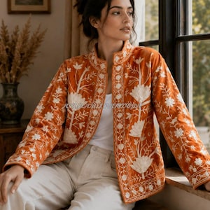 Könnte beinhalten: Eine orangefarbene Jacke mit weißer Blumenstickerei, Stehkragen und langen Ärmeln. Das Kleidungsstück wird über einem weißen Oberteil und einer hellen Hose getragen. Der Text ASSUHANDICRAFTINDIA ist in der Mitte der Jacke zu sehen.