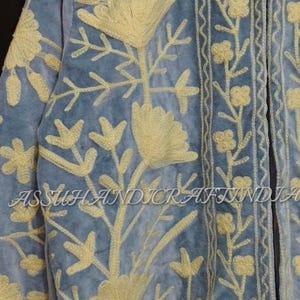 May include: A light blue jacket featuring intricate cream-colored floral embroidery throughout the front and collar. The garment displays the text ASSUHANDICRAFTINDIA across the center. The design includes detailed botanical patterns and borders.