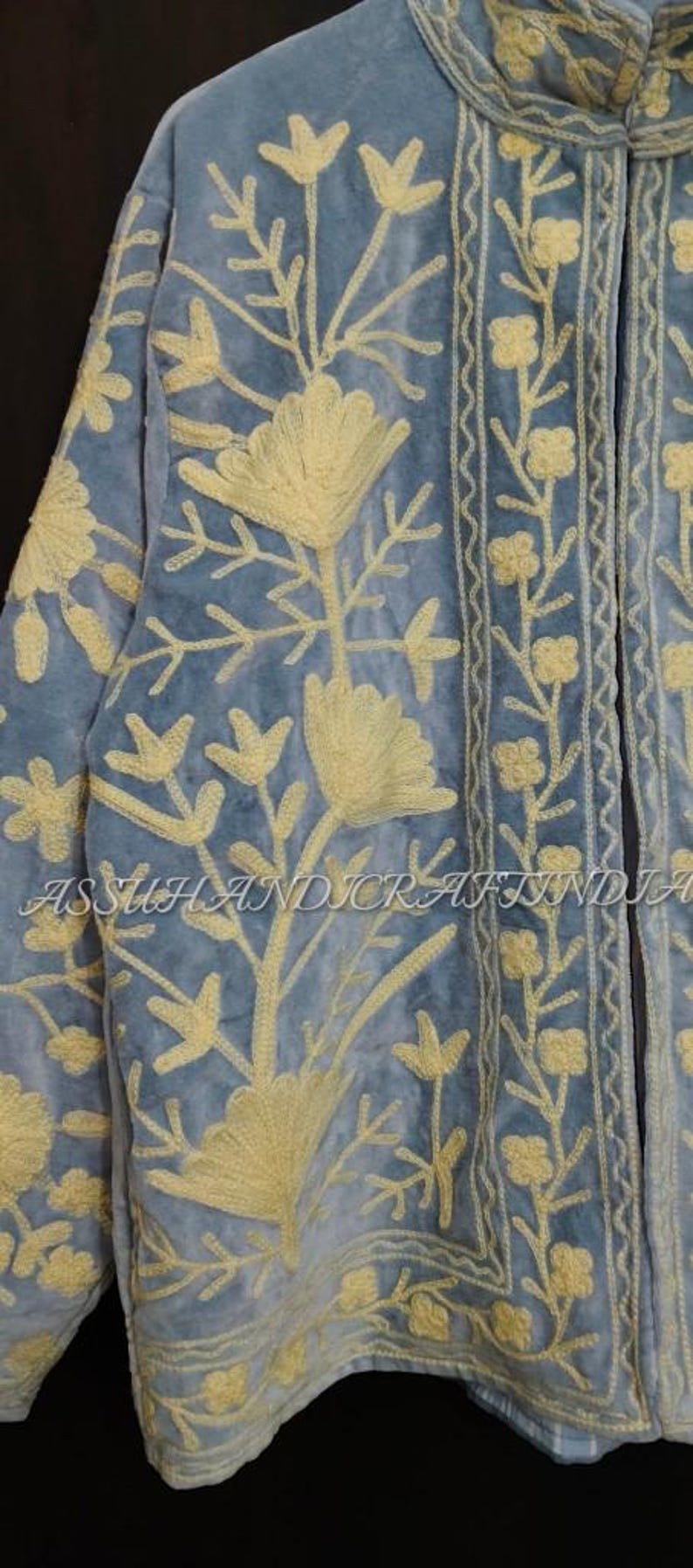 May include: A light blue jacket featuring intricate cream-colored floral embroidery throughout the front and collar. The garment displays the text ASSUHANDICRAFTINDIA across the center. The design includes detailed botanical patterns and borders.