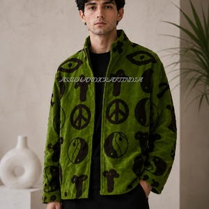 Può includere: Una giacca verde brillante con cerniera caratterizzata da un motivo ripetuto di simboli marrone scuro, tra cui simboli della pace, icone yin-yang e forme di occhi. Il testo ASSUHANDICRAFTINDIA è stampato sulla zona del petto del capo.