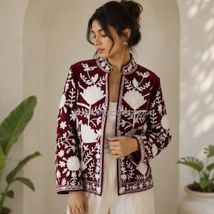 Op de afbeelding: Een diepbordeauxrode jas met een opstaande kraag en lange mouwen, versierd met ingewikkeld wit bloemenborduursel op de voorzijde en de mouwen. Het kledingstuk wordt gedragen over een effen wit topje en een witte broek voor een contrast.