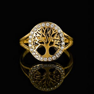 Peut inclure: Bague dorée ornée d'un motif d'arbre de vie, entouré de petits diamants transparents. L'anneau de la bague se divise en deux brins de chaque côté. Le bijou est reflété sur une surface noire.