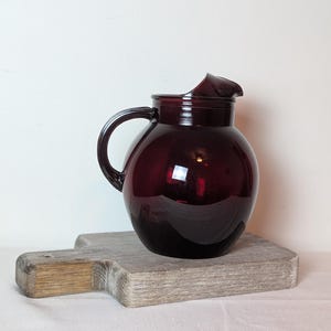 May include: A deep burgundy glass pitcher with a rounded, bulbous body, a wide mouth, and a curved handle on the side. The glass has a smooth, reflective surface that catches light, showing a dark red hue throughout the entire vessel structure.