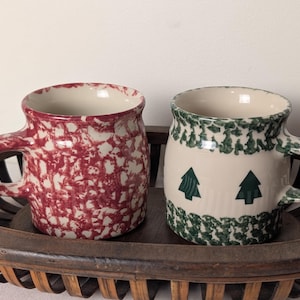 May include: Two ceramic mugs with handles. The left mug features a red and white speckled pattern. The right mug has a cream-colored base with green tree graphics and green speckled bands at the top and bottom rims.