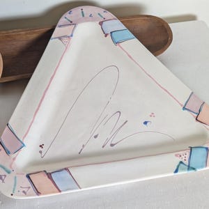 May include: A triangular ceramic serving platter with a white base, featuring a hand-painted border of soft pink, light blue, and peach rectangular accents. The center of the platter displays a thin, dark gray abstract line drawing.