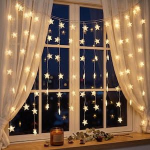 May include: A string of warm white LED star-shaped lights hanging in front of a window with sheer white curtains. The decorative light strands feature multiple vertical drops, each adorned with small, glowing star-shaped bulbs emitting a soft light.