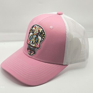 Könnte beinhalten: Eine rosa Baseballkappe mit weißem Netzrücken und einem gestickten Totenkopf-Aufnäher auf der Vorderseite. Der Totenkopf ist mit bunten floralen und geometrischen Mustern in Blau, Gelb, Rot und Schwarz auf dem festen rosa Vorderteil verziert.