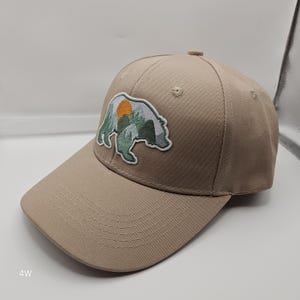 Könnte beinhalten: Eine beigefarbene Baseballkappe mit einem gestickten Aufnäher auf der Vorderseite. Der Aufnäher zeigt die Silhouette eines Bären, gefüllt mit einer Landschaft aus grünen Bergen und einer leuchtend orangefarbenen Sonne auf weißem Grund.