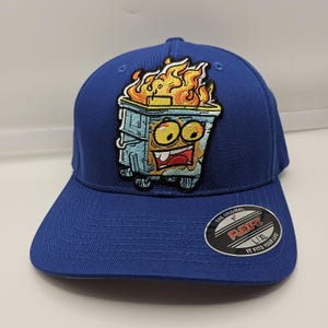 Puede incluir: Una gorra de béisbol azul real con un parche bordado de un contenedor de basura en llamas con ojos grandes y una sonrisa dentuda. La visera incluye una pegatina circular negra y plateada con el texto Flexfit The Original It Fits Your Life.