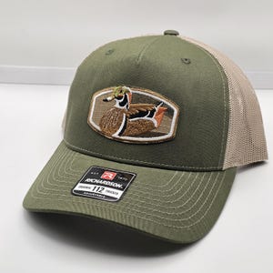Puede incluir: Gorra de camionero verde oliva con parte trasera de malla beige y visera curva. La parte delantera presenta un parche bordado de un pato. La etiqueta indica Richardson 112 Trucker.