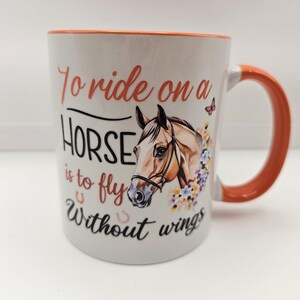 May include: White ceramic mug with an orange handle and rim. The mug features a horse head illustration with floral accents and the text "To ride on a HORSE is to fly Without wings."