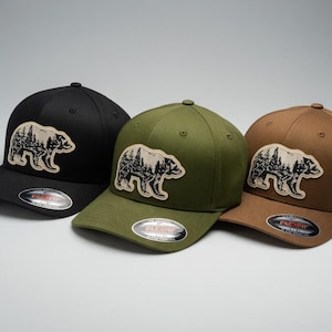 Peut inclure: Trois casquettes de baseball noires, vert olive et marron. Chaque casquette arbore un écusson d'ours et de forêt à l'avant. Les casquettes ont une visière incurvée et un design ajusté. Elles sont présentées sur une surface gris clair.