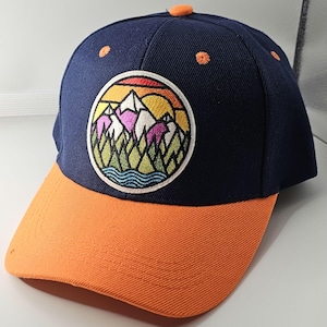 May include: A navy blue baseball cap with an orange peak. The cap has an embroidered patch with a mountain and sunset design. The cap has orange stitching and a small orange button on top.