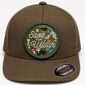 Może przedstawiać: Ciemnozielona oliwkowa czapka bejsbolowa z okrągłą naszywką z napisem "Save Our Wildlife". Naszywka przedstawia czarnego ptaka, kwiaty, grzyb, borsuka i rybę. Czapka ma zakrzywiony daszek.