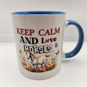 May include: White ceramic mug with a blue interior and handle. The mug features the text "KEEP CALM AND Love HORSES" in red and black. A graphic of a horse in a field of flowers and butterflies is also on the mug.