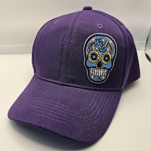 May include: A purple baseball cap with a decorative patch featuring a colorful sugar skull design. The patch includes a blue rose, yellow accents, and a white toothy grin. The cap has a curved brim and a button on top.