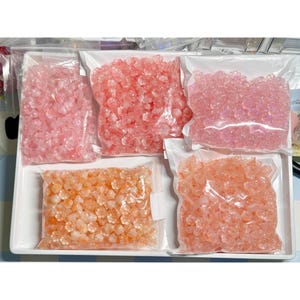 May include: A white tray holds five clear plastic bags filled with small, flower-shaped beads in shades of pink and orange. The beads appear to be made of a translucent material, and the bags are sealed with a zipper closure.