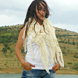 May include: A woman wearing a white crocheted shawl with fringe, a white tank top, and blue jeans. She is standing in front of a grassy hillside.