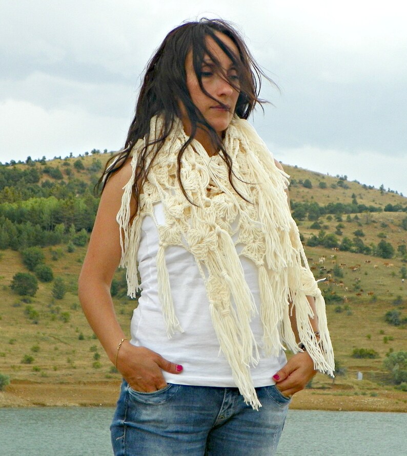 May include: A woman wearing a white crocheted shawl with fringe, a white tank top, and blue jeans. She is standing in front of a grassy hillside.