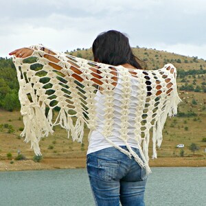 May include: A white crochet shawl with fringe, worn by a person with long dark hair. The shawl has a lacy pattern and is draped over the person's shoulders.