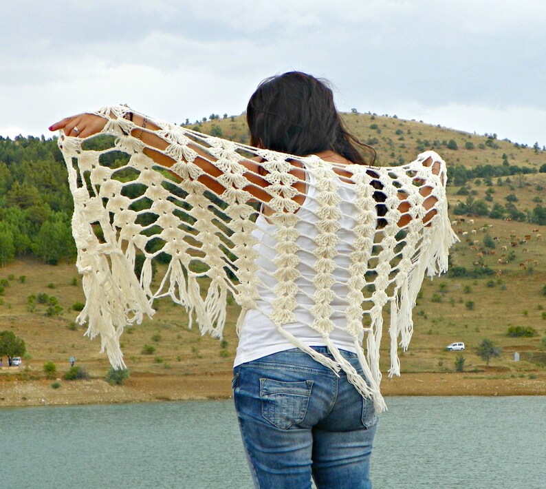 May include: A white crochet shawl with fringe, worn by a person with long dark hair. The shawl has a lacy pattern and is draped over the person's shoulders.