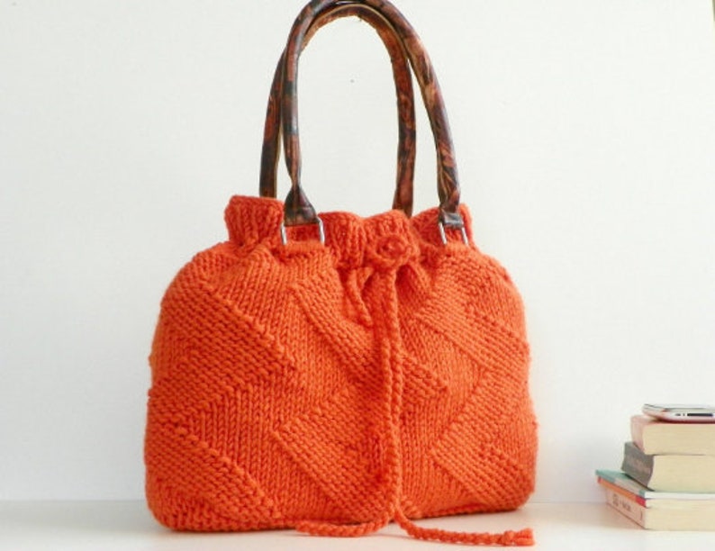 May include: Orange knitted tote bag with leather handles. The bag has a drawstring closure and a diamond pattern.