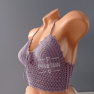 Pu&ograve; includere: Un top halter in crochet viola con un dettaglio di nodo sul davanti. Il top ha un delicato motivo a pizzo ed &egrave; mostrato su un manichino. Il testo "@N2LDESIGNN" &egrave; visibile sul top.