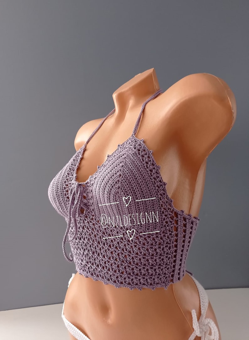 Pu&ograve; includere: Un top halter in crochet viola con un dettaglio di nodo sul davanti. Il top ha un delicato motivo a pizzo ed &egrave; mostrato su un manichino. Il testo "@N2LDESIGNN" &egrave; visibile sul top.