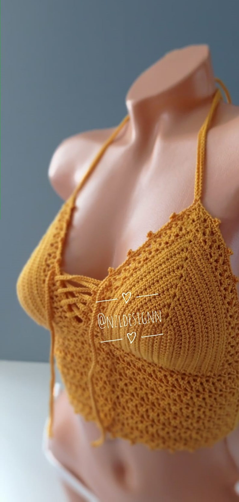 Pu&ograve; includere: Un top halter in crochet giallo senape con un dettaglio di pizzo sul davanti. Il top presenta un delicato motivo a crochet ed &egrave; realizzato con un filato morbido e leggero. Il testo "@NZLDESIGNN" &egrave; visibile sul top.