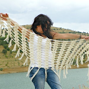 May include: A white crocheted shawl with fringe, worn by a person with long dark hair. The shawl is draped over the person's shoulders and arms, and the fringe is hanging down the sides.