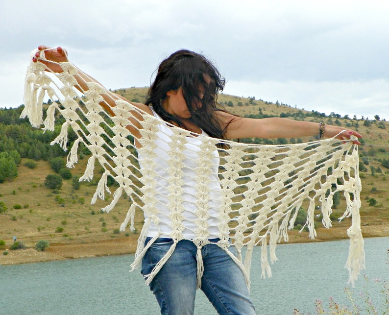 May include: A white crocheted shawl with fringe, worn by a person with long dark hair. The shawl is draped over the person's shoulders and arms, and the fringe is hanging down the sides.