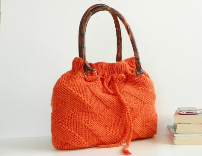 May include: Orange knitted tote bag with brown handles. The bag has a drawstring closure and a diamond pattern.
