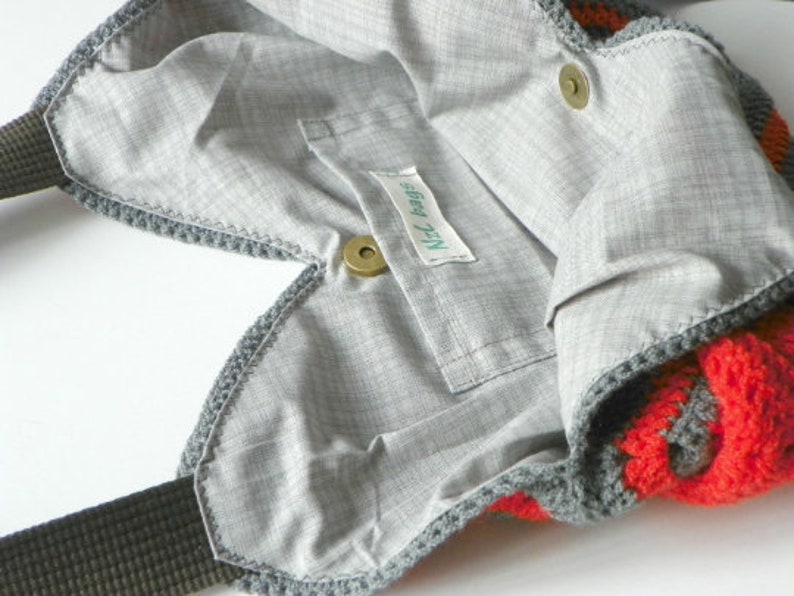 May include: A gray crocheted tote bag with a red and gray striped pattern. The bag has a zipper closure and a small pocket on the inside. The bag has a label that says "Nici Bags".
