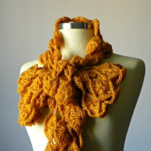 May include: A crocheted scarf in a deep mustard yellow color. The scarf is made of a textured yarn and has a ruffled design.