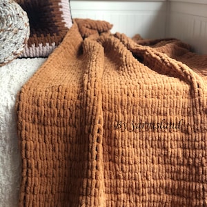 May include: A brown knitted throw blanket with a fluffy texture. The blanket is folded and draped over a white bed with pillows.