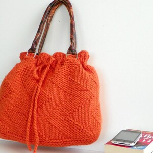 May include: Orange knitted tote bag with brown leather handles. The bag has a drawstring closure and a diamond pattern.