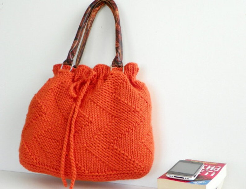 May include: Orange knitted tote bag with brown leather handles. The bag has a drawstring closure and a diamond pattern.
