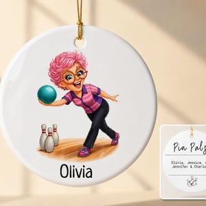 May include: A round white ceramic ornament featuring a cartoon illustration of a person with pink hair and glasses bowling a teal ball toward three pins. The name Olivia is printed below the illustration. A gold string is attached at the top.