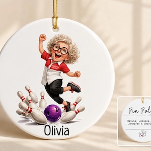 May include: A round white ceramic ornament featuring a cartoon illustration of a curly-haired person in a red and white shirt labeled Bowling Queen, jumping over a purple bowling ball and white pins. The name Olivia is printed below the illustration.