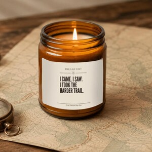 May include: A lit candle in an amber glass jar with a white label featuring the text: THE LILY EDIT, I CAME. I SAW. I TOOK THE HARDER TRAIL. 9 oz | Natural Soy Wax. The candle is positioned on a vintage map next to a brass compass.