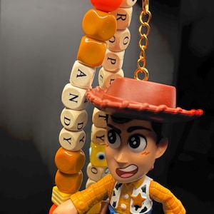 May include: A Woody keyring featuring a miniature figurine of the character with a cowboy hat, plaid shirt, and blue trousers. The keyring includes a beaded charm spelling out "ANDY" and "PROPERTY". The charm is attached to a gold-coloured clasp.