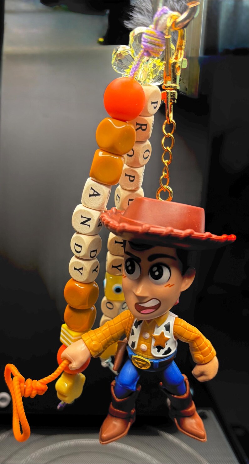 May include: A Woody keyring featuring a miniature figurine of the character with a cowboy hat, plaid shirt, and blue trousers. The keyring includes a beaded charm spelling out "ANDY" and "PROPERTY". The charm is attached to a gold-coloured clasp.